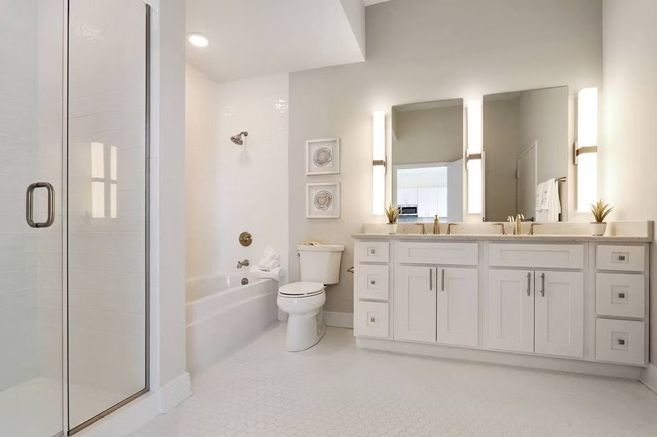 White Shaker bathroom with double vanity, lighted mirrors, gold hardware and glass shower